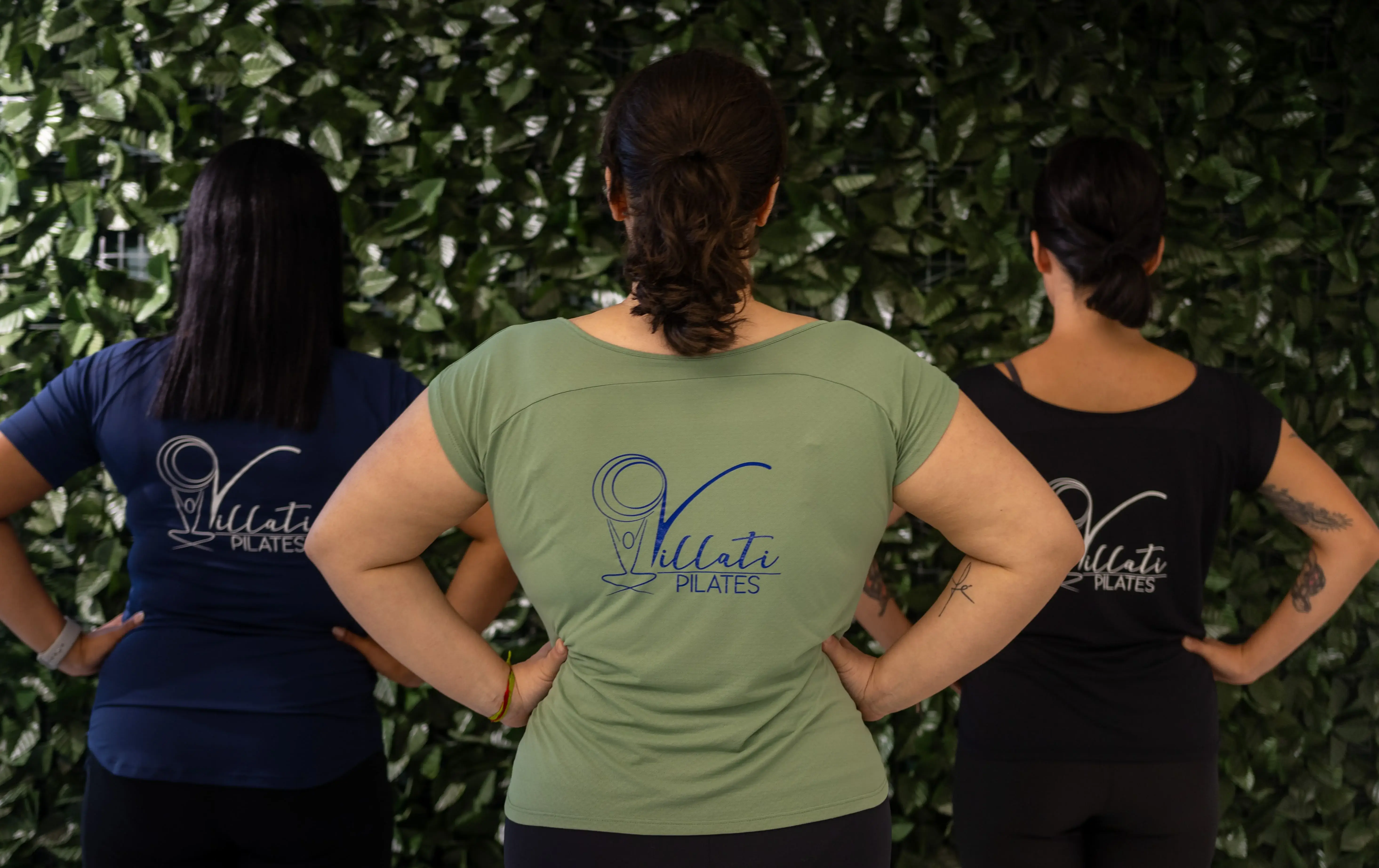 Três alunas de costas, vestindo camisetas da Villati Pilates, em frente a uma parede de folhagens